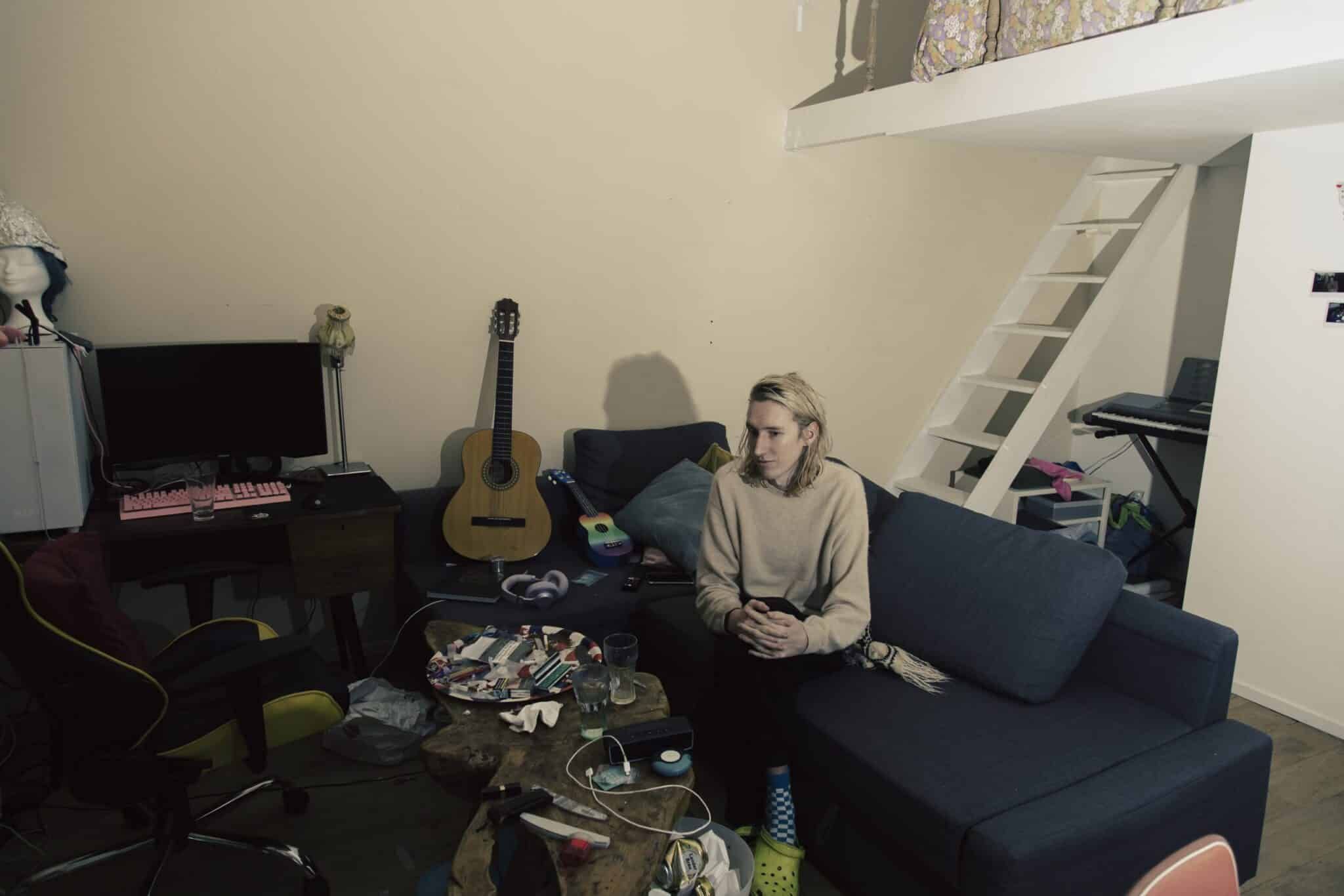 Autonoom persoon zit op een bank in een rommelige kamer met gitaar en bureau. Documentaire foto. Joep van Drunen Photography